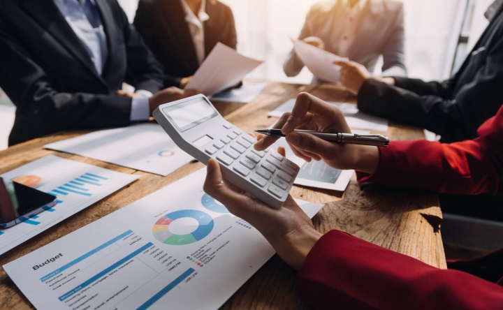 **Alt text:** A person holds a calculator and pen while reviewing financial charts on a desk, with colleagues in the background discussing documents during a business meeting.