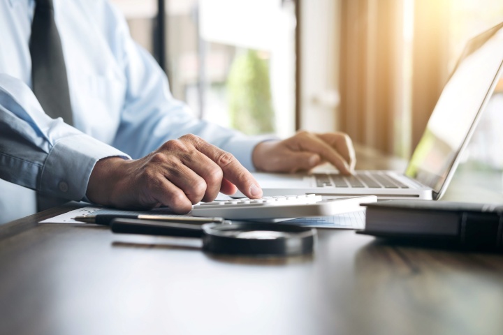 **Alt text:**A business professional uses a calculator beside a laptop at a desk, with documents and office items nearby, suggesting financial analysis or accounting work.
