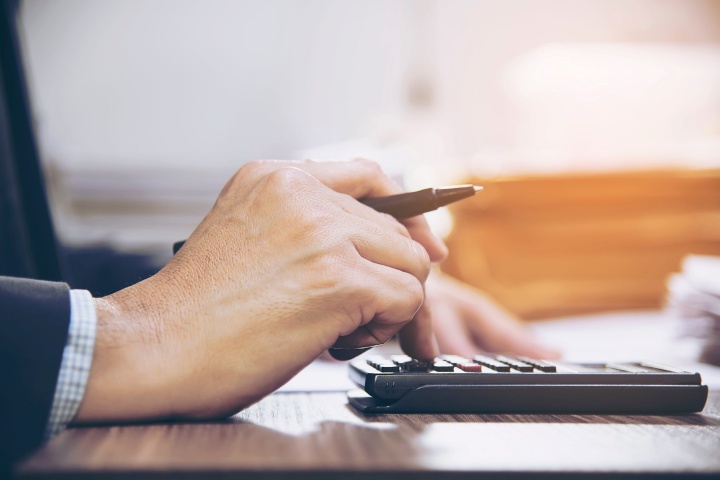 **Alt text:**A close-up of a person using a calculator while holding a pen, working on paperwork at a desk in a softly lit office setting.
