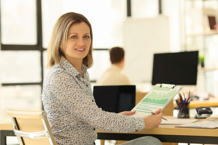 **Alt text:**A woman sitting at a desk in a bright office holds a clipboard with a chart, smiling toward the camera, with a computer and coworker in the background.