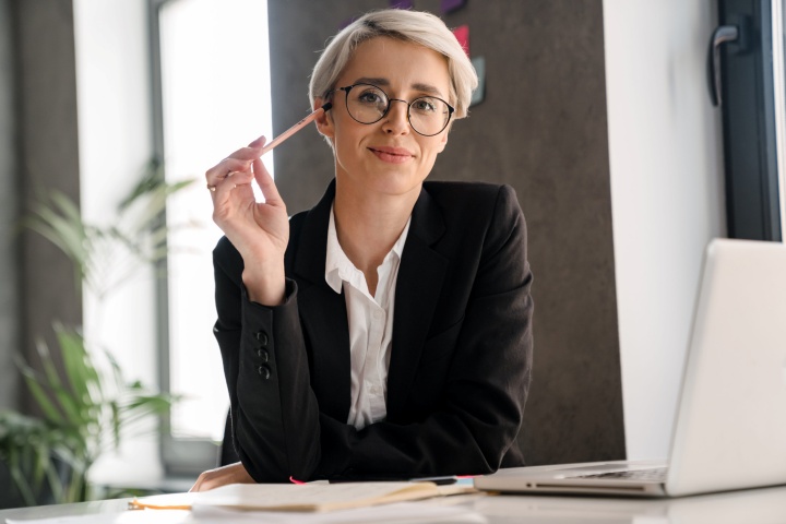 **Alt text:**A professional woman wearing glasses and a black blazer sits at a desk, holding a pencil and smiling confidently toward the camera, with a laptop and notebook in a modern office setting.

