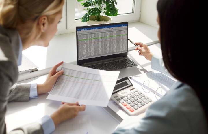 **Alt text:**Two colleagues review financial data together at a desk, comparing printed spreadsheets while looking at a laptop displaying a detailed spreadsheet, with a calculator nearby.